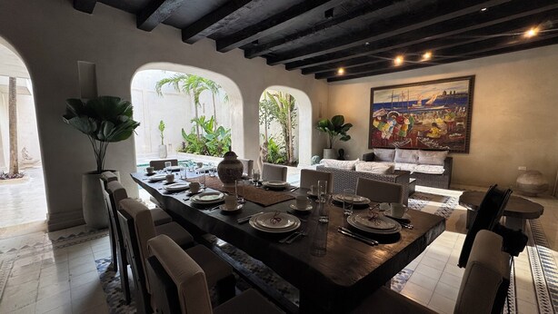 A dining area with a large table set for a meal, surrounded by chairs. There are plants and a painting on the walls. - Casa Bovedas Collection by Ritzy Group (Cartagena de Indias)