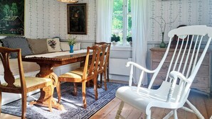 A dining area with a wooden table, chairs, and a white rocking chair. There is a window with curtains, a painting on the wall, and a vase with flowers on the table. - Pet friendly home in Malmbäck with WiFi (Malmbäck)