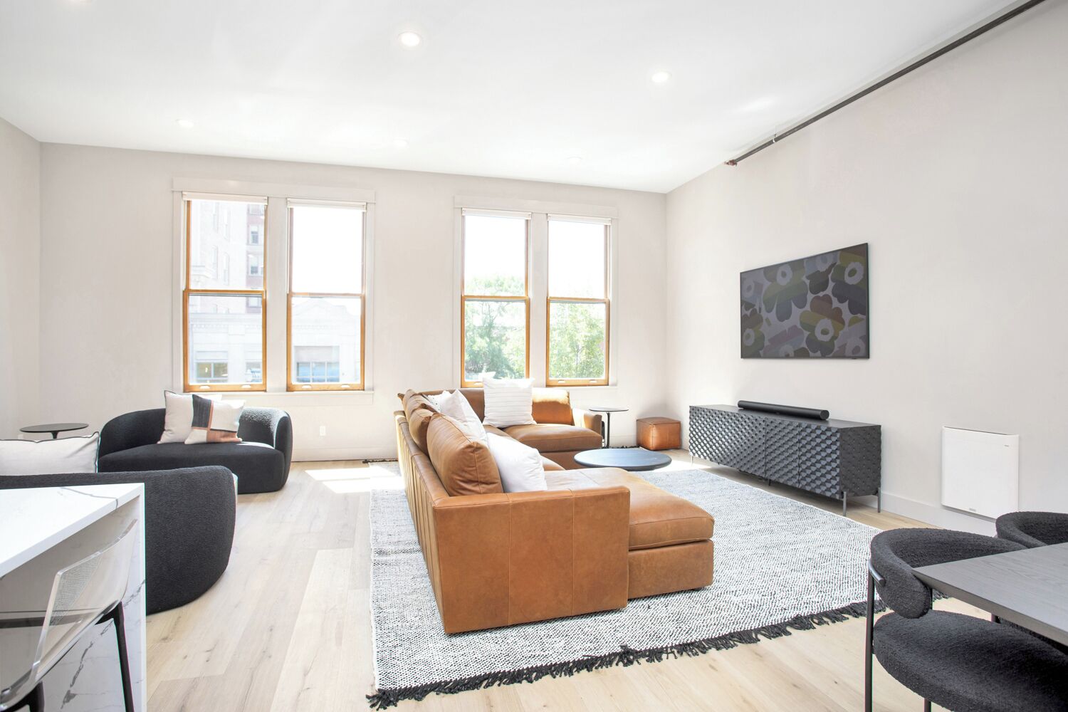 A modern living room with a brown sofa, a round coffee table, and a grey cabinet. There are two windows providing natural light and a painting on the wall.