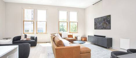 A modern living room with a brown sofa, a round coffee table, and a grey cabinet. There are two windows providing natural light and a painting on the wall.