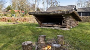 Ein rustikales Holzgebäude mit geneigtem Dach, vermutlich eine Hütte oder eine kleine Schutzhütte, umgeben von einer Wiese mit einem Feuer davor.