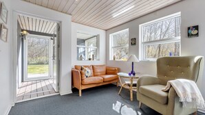 Ein gemütliches Wohnzimmer mit einem Ledersofa, einem beigefarbenen Sessel und einem kleinen runden Tisch mit einer Lampe. Große Fenster sorgen für natürliches Licht und Blick ins Freie.