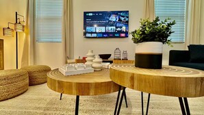 Un salon moderne avec deux petites tables rondes en bois, une grande plante en pot et un téléviseur à écran plat fixé au mur, affichant l’interface de YouTube TV.