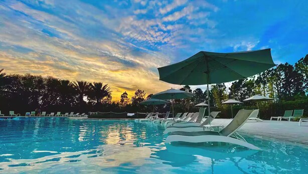 Une piscine entourée d’arbres, avec des chaises longues et des parasols, le tout baigné par la lumière d’un coucher de soleil.