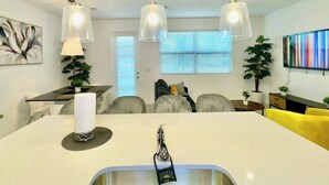 A modern kitchen with a central island, pendant lights, a dining area with a table and chairs, and a living area with a sofa and a TV.