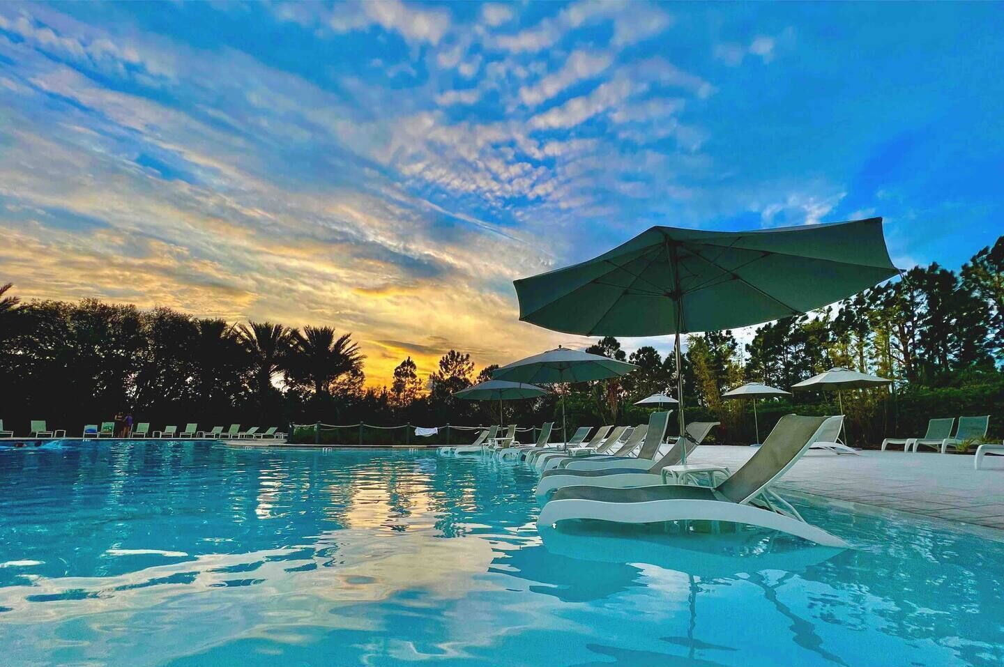 Una piscina con sillas de descanso y sombrillas, rodeada de árboles y un cielo al atardecer.