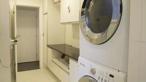 A laundry room with a front-loading washing machine and dryer, a small shelf with items, and a door leading to another room. - ROCK IN RIO 2026 PROMO Sep 5th to 14th
Best Location Best Living in Rio. (Rio de Janeiro)