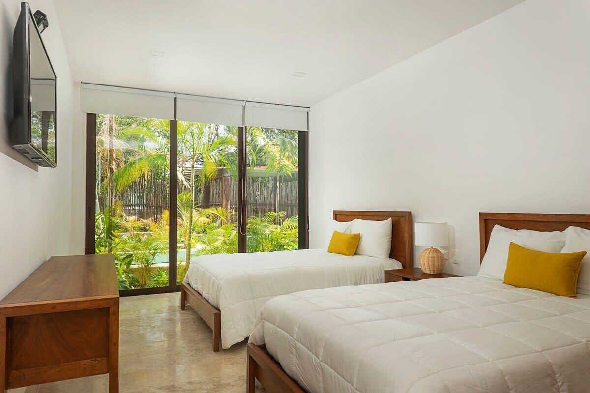 A hotel room with two beds, a wooden nightstand, a TV mounted on the wall, and a large sliding glass door leading to a tropical garden.