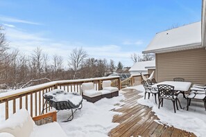 Une terrasse en bois, meublée pour l’extérieur et recouverte de neige, avec une balustrade et une vue sur des arbres et un bâtiment au loin.