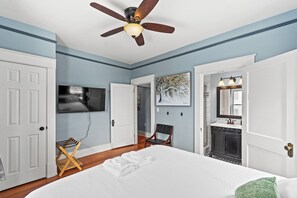 Une chambre à coucher comprenant un lit, un ventilateur de plafond, une télévision, une chaise, un tableau, ainsi qu’une salle de bain avec un lavabo et un miroir.