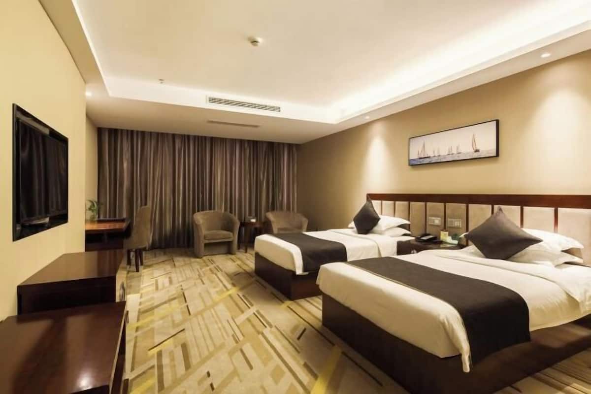 A hotel room with two beds, a desk, a chair, a large window with curtains, and a painting on the wall.