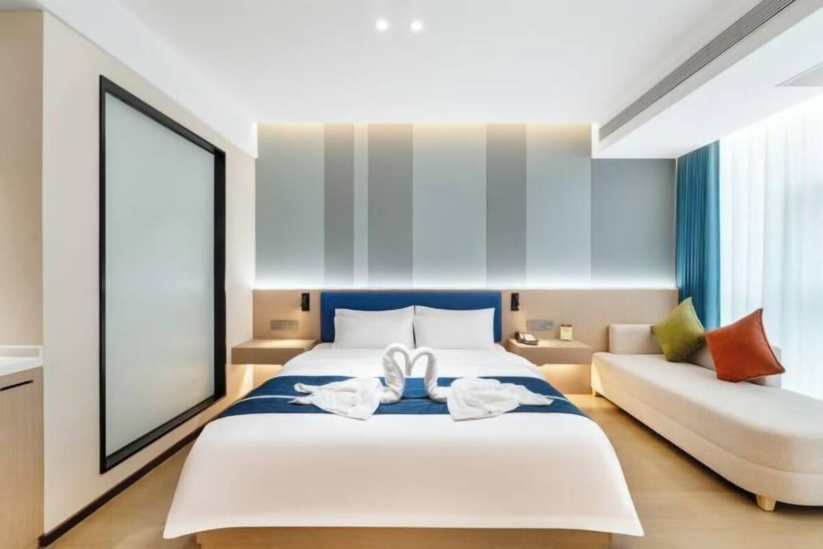 A modern hotel room with a large bed, a sofa, and a large window with curtains.
