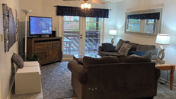 A living room with a sofa, television, ceiling fan, and a view of the outside through a door.