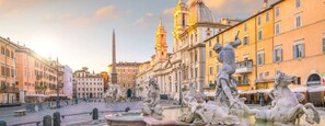 Una plaza histórica con estatuas clásicas y fuentes, rodeada de edificios tradicionales.