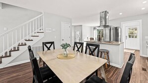 Une cuisine moderne dotée d’une hotte en acier inoxydable, un coin repas avec une table en bois et des chaises noires, et un escalier avec des rampes blanches.