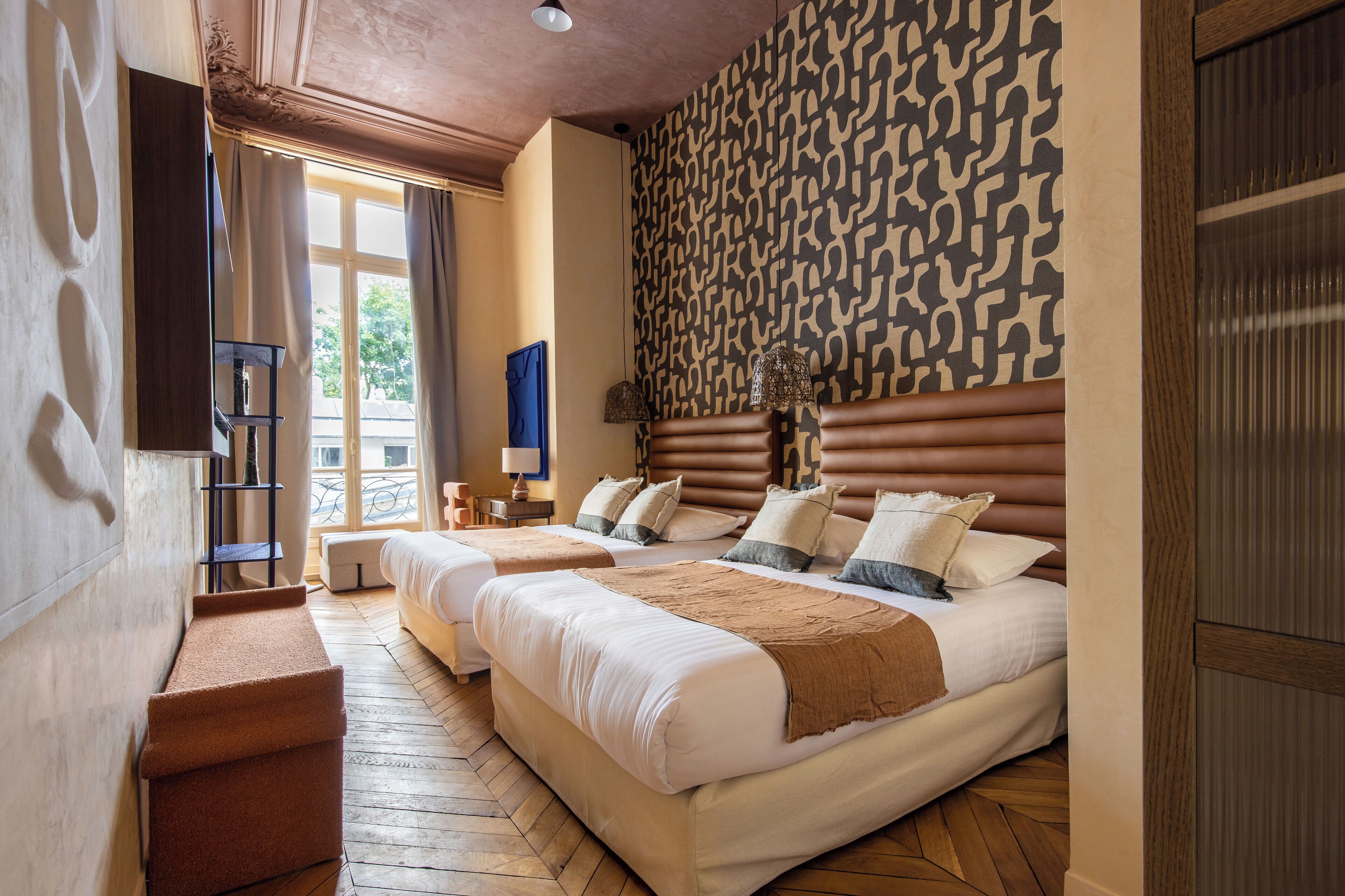 A hotel room with two beds, a patterned wall, a wooden floor, and a view through a window.