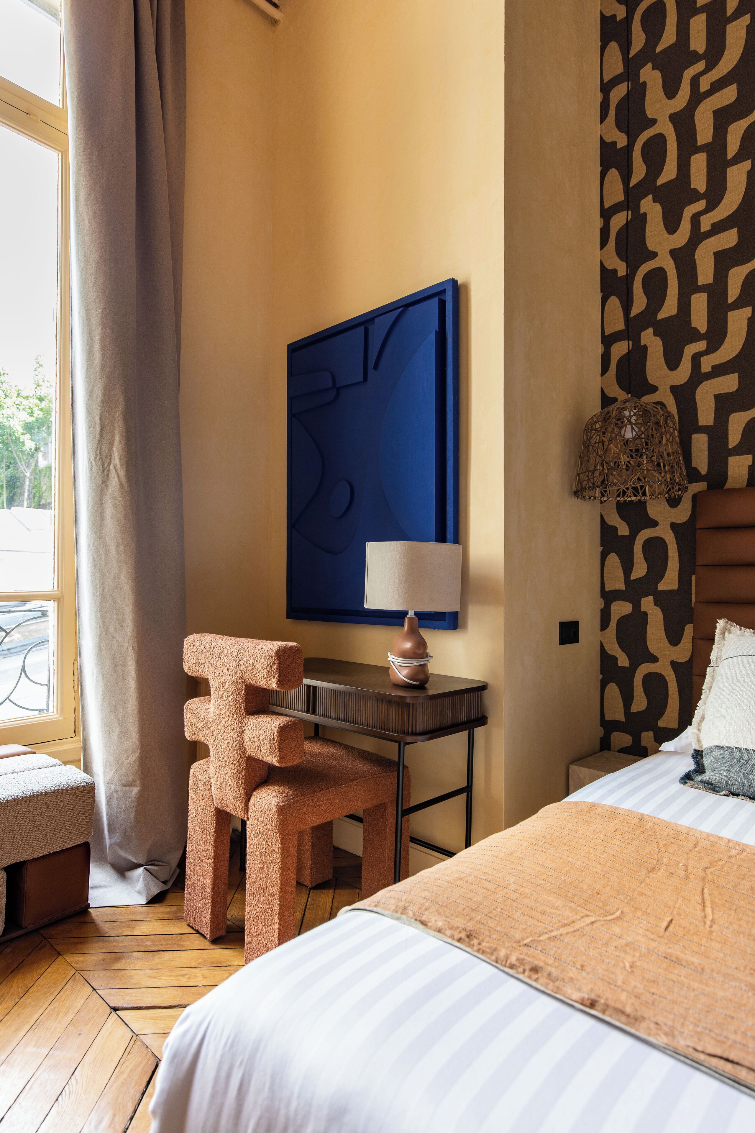 A hotel room with a bed, a chair, a table with a lamp, a wall art piece, and a patterned wall.