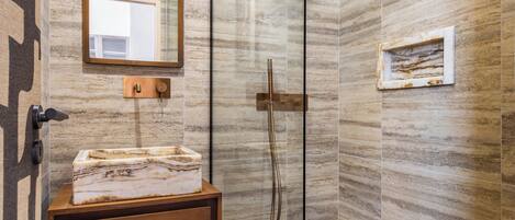 Un baño moderno con ducha envuelta en vidrio, tocador de madera con lavamanos de mármol y un espejo sobre el tocador.