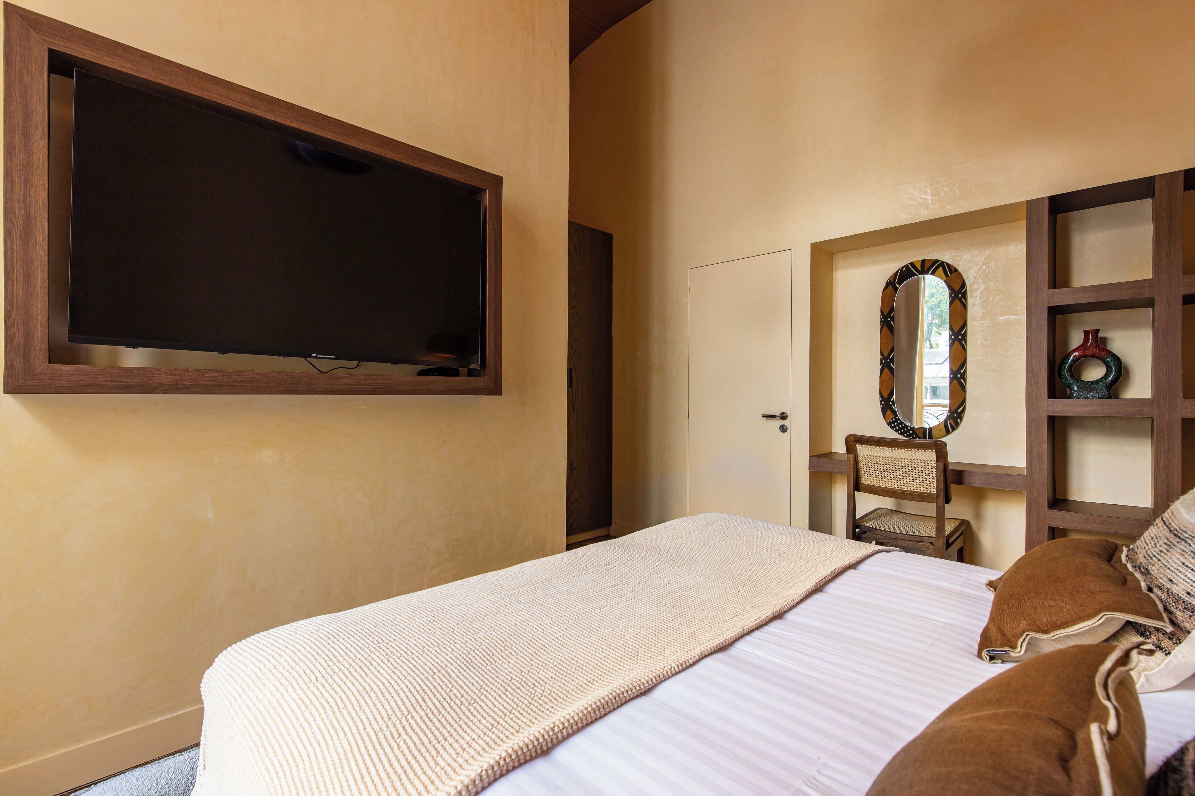 Een slaapkamer met een groot bed, een aan de muur bevestigde televisie, een spiegel en een klein houten bureau met een stoel.