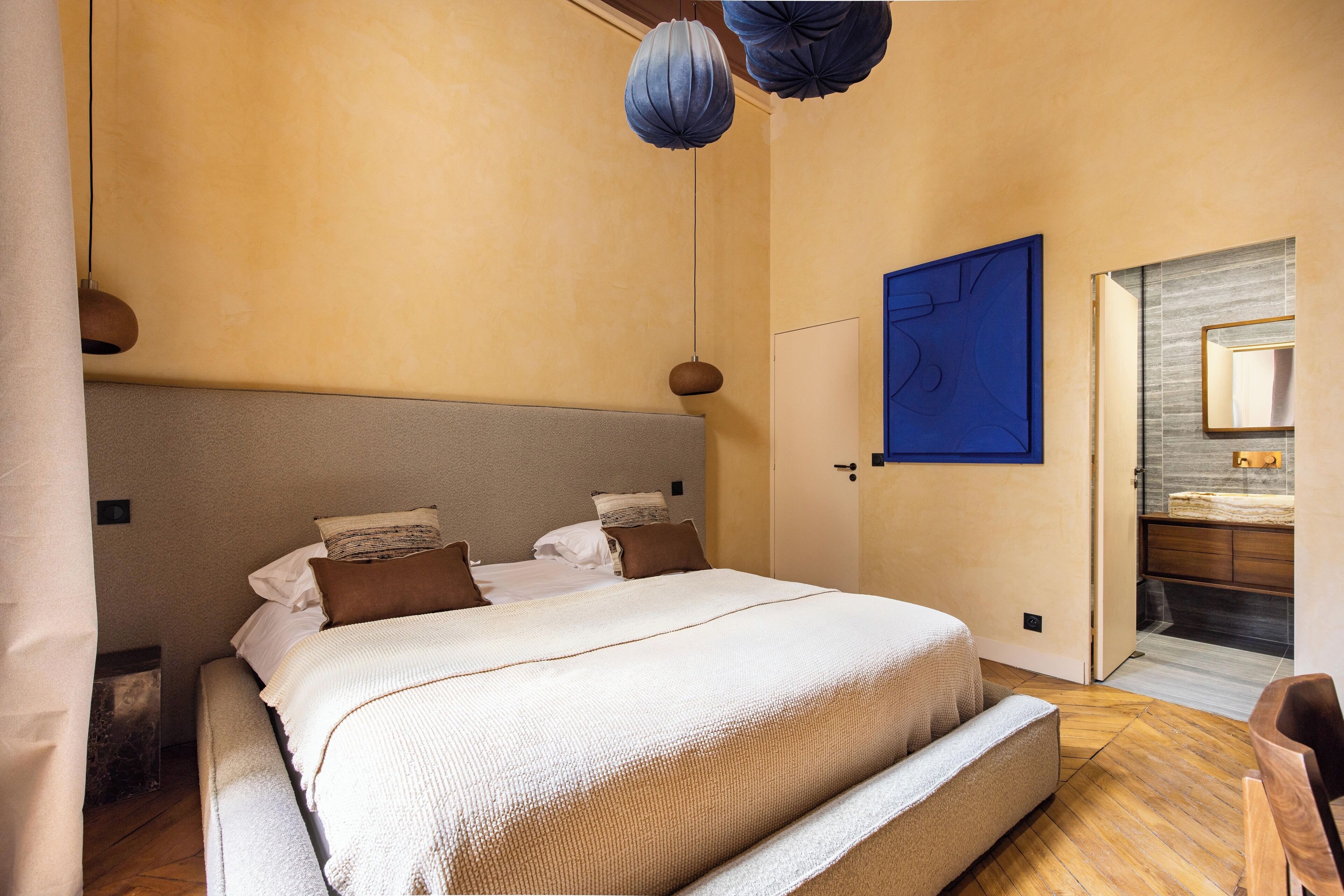 A bedroom with a large bed, a wooden floor, a blue artwork on the wall, and a bathroom with a mirror and sink.
