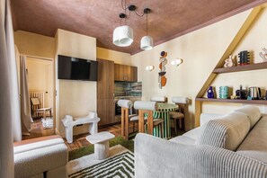 A modern living room with a sofa, a coffee table, and a TV mounted on the wall. There is a kitchen area with wooden cabinets and a dining table with chairs.