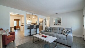 Un salon moderne avec un canapé gris, un fauteuil rouge et une table basse blanche. On aperçoit une cuisine à l’arrière-plan, avec un réfrigérateur et un coin repas.