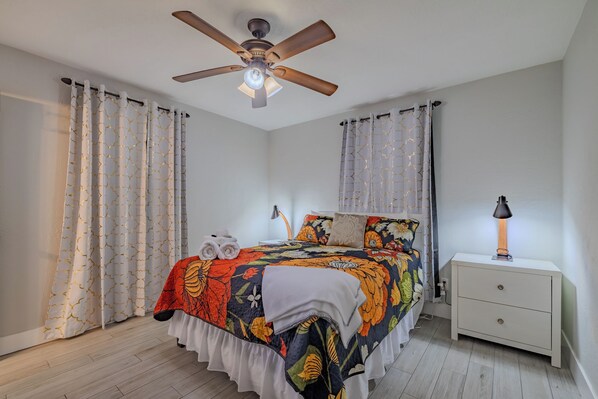 Une chambre à coucher avec un lit, des rideaux, un ventilateur de plafond, une lampe de chevet et une table de nuit.