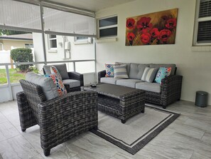 A covered patio area with wicker furniture, a gray sofa with colorful pillows, and a painting of red flowers on the wall.