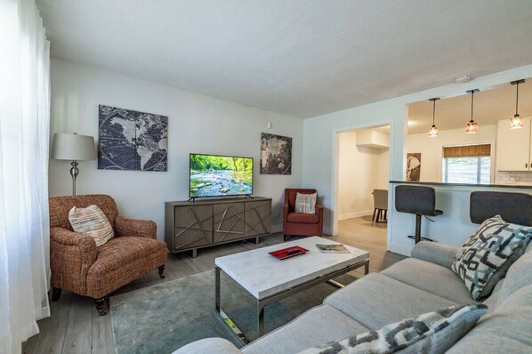 Un salon moderne avec un canapé, un fauteuil et une table basse. On y trouve un téléviseur posé sur un meuble, et des œuvres d’art accrochées aux murs.