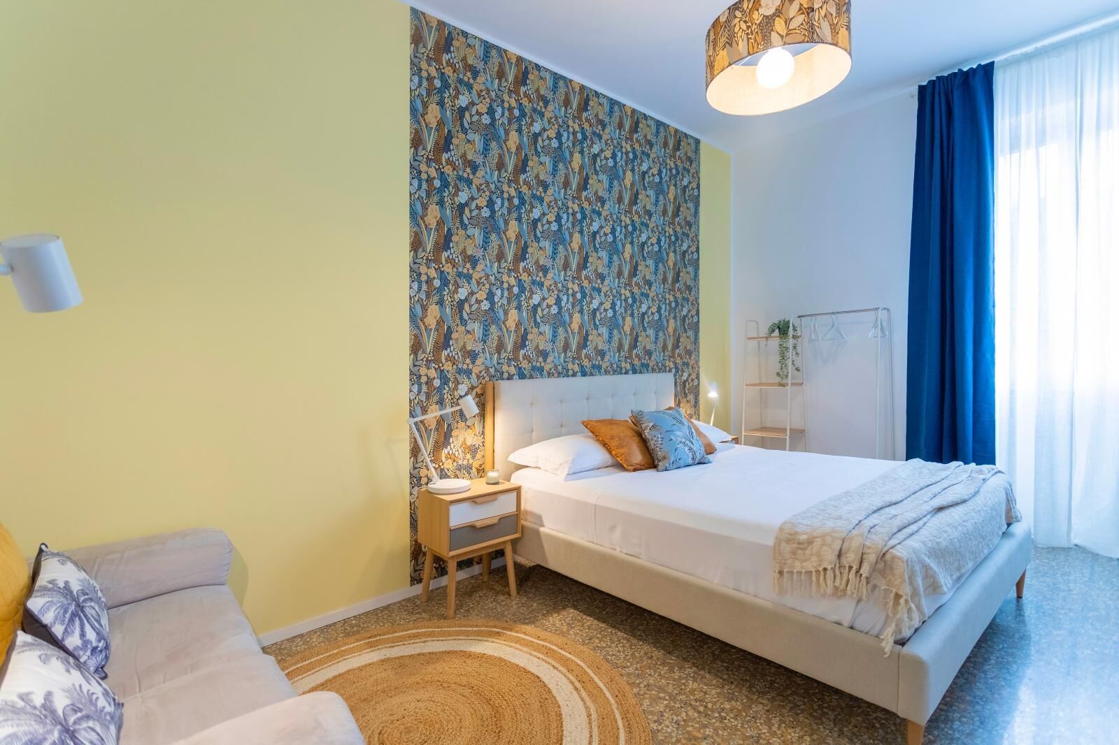 A hotel room with a large bed, a bedside table with a lamp, a nightstand, a sofa, and a patterned wall.