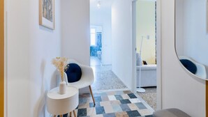 A modern hallway with a checkered floor, a white chair with a blue cushion, a small table with a vase, and a mirror on the wall. - Flora Cozy Flat close to Metro (ROMA)