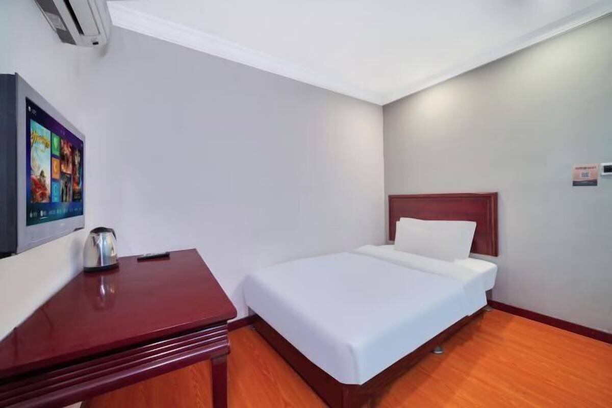 A hotel room with a bed, a wooden desk, a television mounted on the wall, and a small table with a kettle.