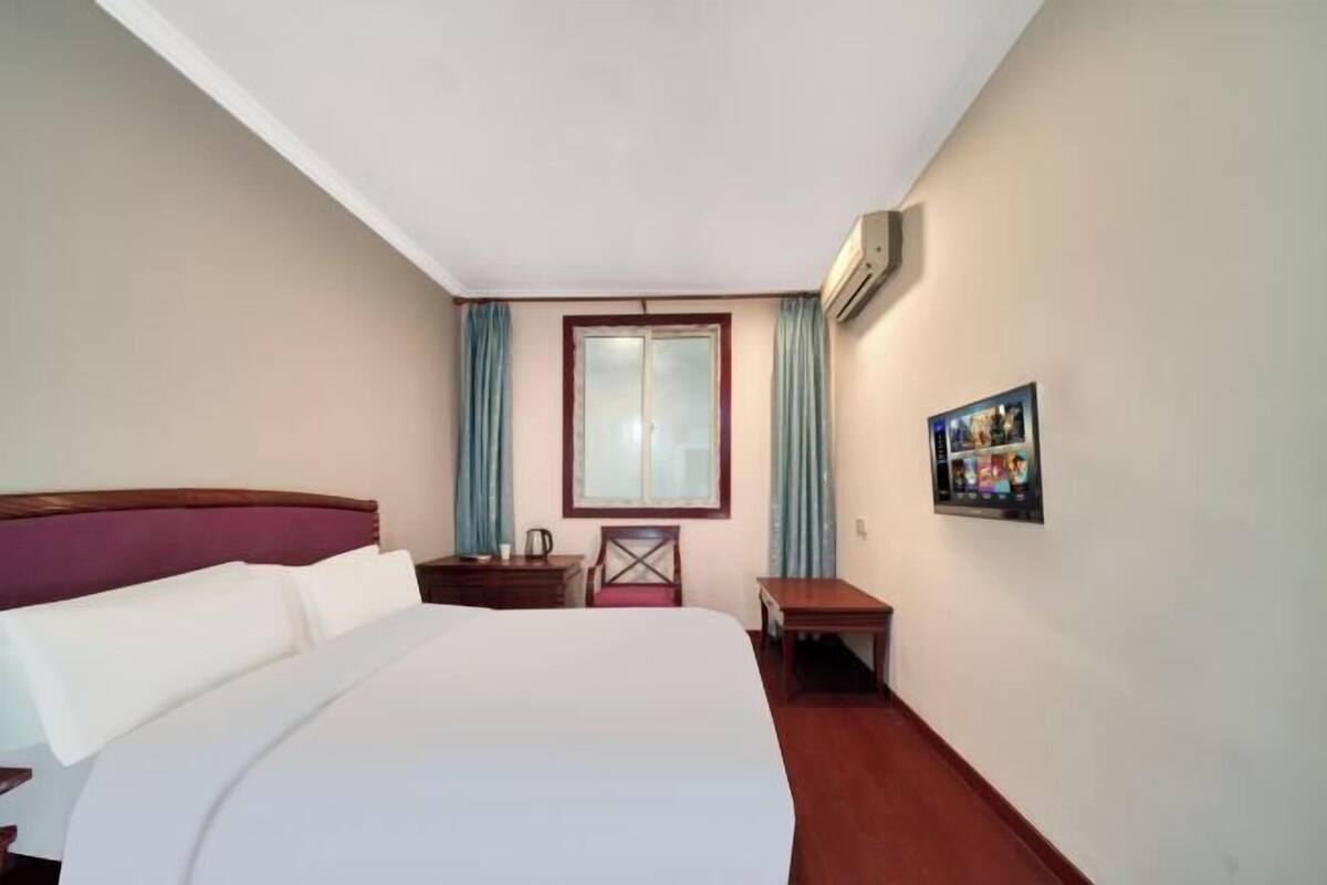 A hotel room with a large bed, a wooden headboard, a nightstand, a small table, a chair, a window with curtains, an air conditioning unit, and a framed picture on the wall.