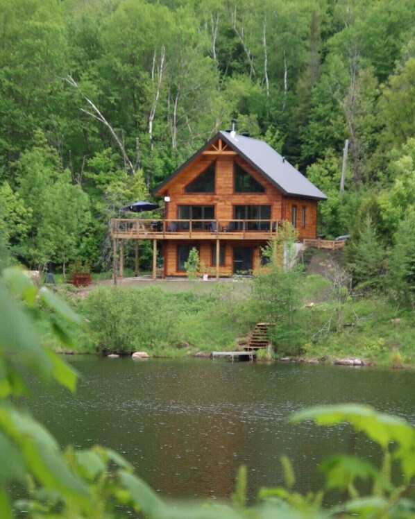 A wooden cabin with a balcony overlooking a lake, surrounded by dense forest.