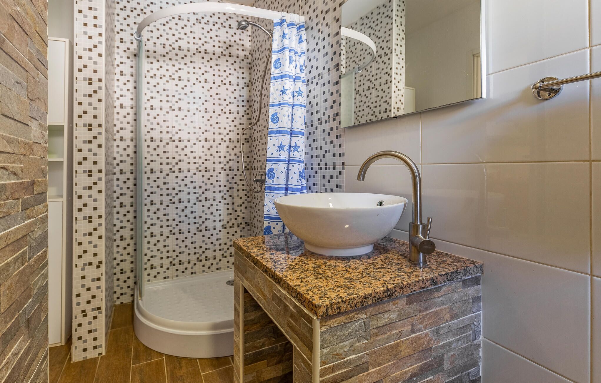 A bathroom with a white sink, a shower with a mosaic tile wall, and a stone countertop.