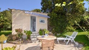 A small, single-story building with a tiled patio, a wooden table, two white chairs, and a blue and white striped awning. - Gorgeous home in OLETTA (OLETTA)