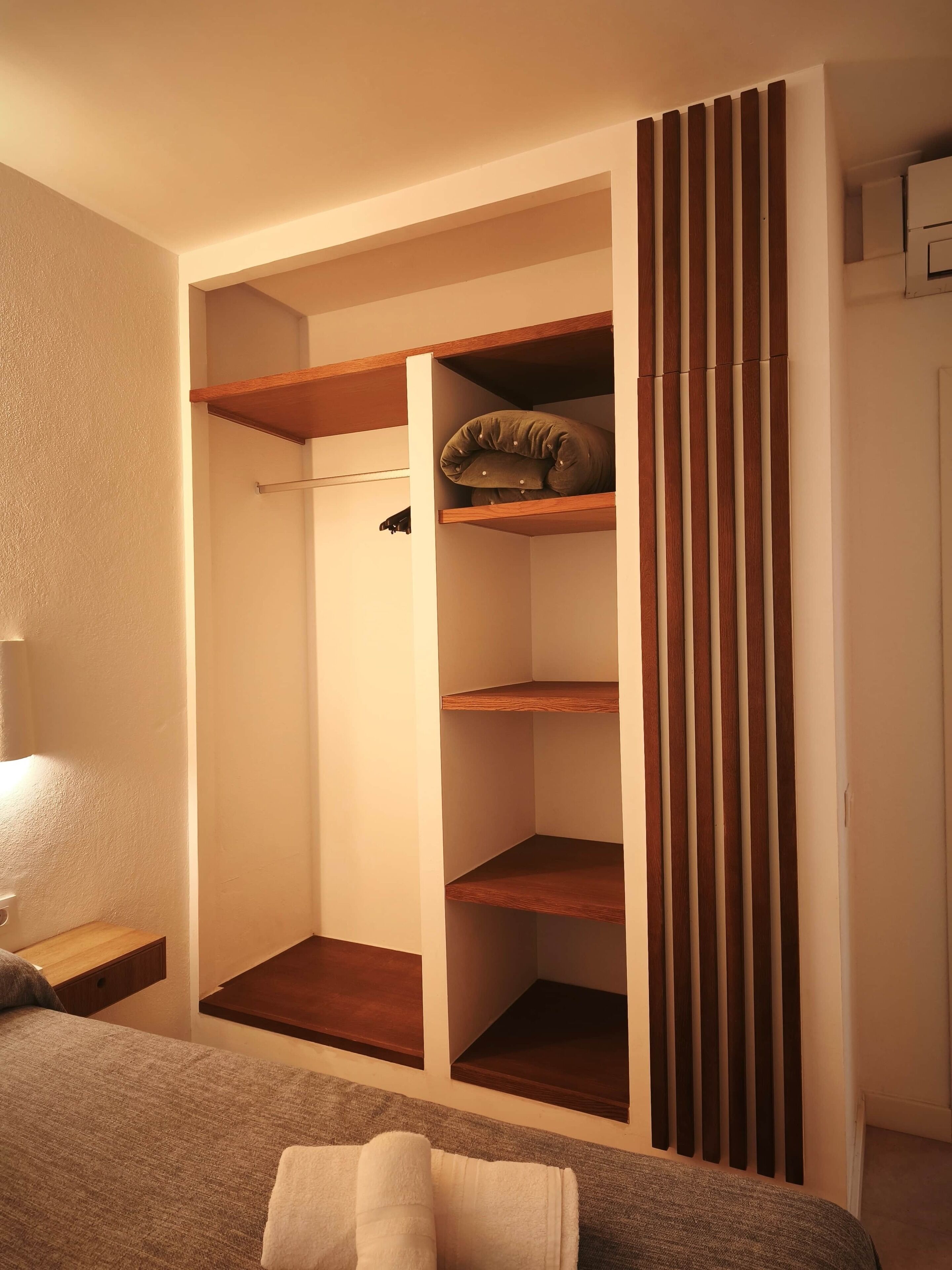 Un dormitorio con una cama, una mesita de noche, un armario con estantes y una cortina.