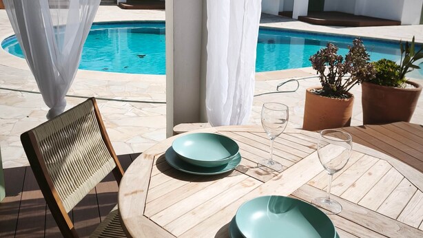 A wooden table with a chair, a glass, and a plate set for dining, overlooking a swimming pool with a white building in the background.