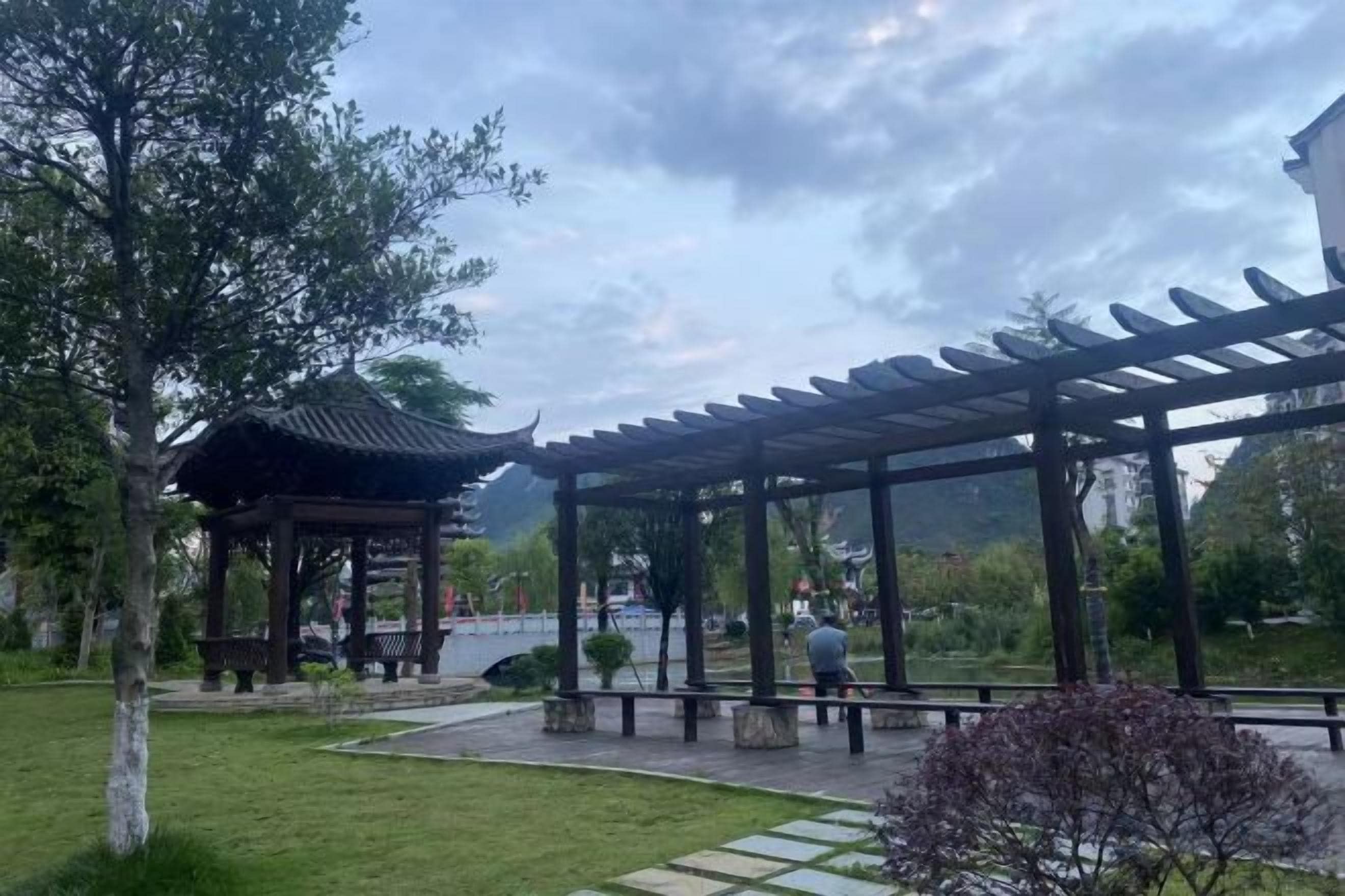 Un mirador con techo, una banca y un sendero en un parque con árboles y un cuerpo de agua al fondo.