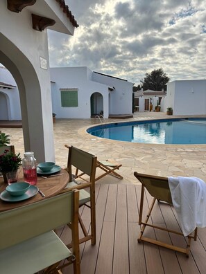 Un espace extérieur avec une piscine, une terrasse en bois, une table avec des chaises et une vue sur des bâtiments blancs en arrière-plan.