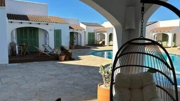 Un espace piscine agrémenté d’un fauteuil suspendu, de plantes en pot et d’un bâtiment doté d’arcades et d’un toit en tuiles.
