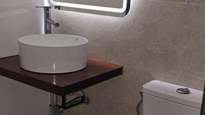 Une salle de bain moderne avec un lavabo blanc, un miroir doté d’un éclairage intégré, des toilettes et une petite étagère avec une poubelle.