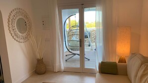Ein Zimmer mit einer Couch, einem Holztisch, einem Ziertegelspiegel, einer Lampe und Blick durch eine Glastür auf einen Außenbereich mit einem Sessel und einem Tisch.