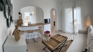 Ein modernes Wohnzimmer mit einem weißen Sofa, einem Holztisch und einer Küchenzeile mit Bar und Hockern.