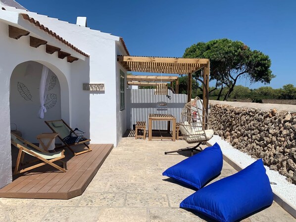 A sunlit outdoor seating area with wooden loungers, a swing chair, and blue cushions.