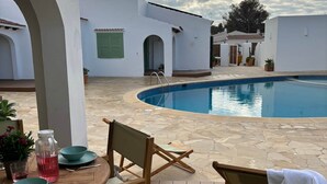 Zona de piscina exterior con terraza de madera, una mesa con sillas y vistas a edificios blancos al fondo.