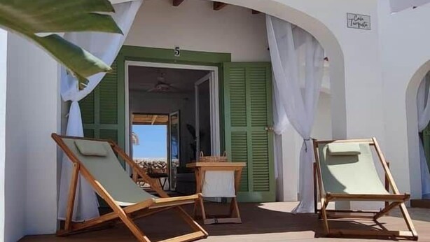 Un espacio al aire libre techado con sillones de madera y una pequeña mesa, un edificio blanco con contraventanas verdes, y un cielo despejado.