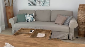 Ett vardagsrum med en beige soffa, ett soffbord i trä, en vit takfläkt, en växt som hänger på väggen och en flätad korg.