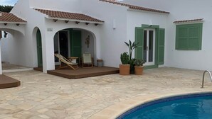 Ett område vid poolen med en stenbelagd uteplats, en pool och en byggnad med gröna fönsterluckor och ett tak täckt med tegelpannor.