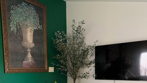 A room with a green sofa, a small ottoman, a television, a framed painting, and a potted plant. - Near O’Hare, King Bed, Spacious, Great Backyard, WiFi, AC (Wood Dale)